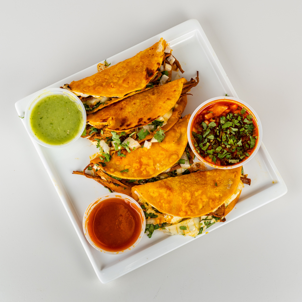 Four golden-brown birria tacos with cilantro and onions, served with three dipping sauces on a white plate.