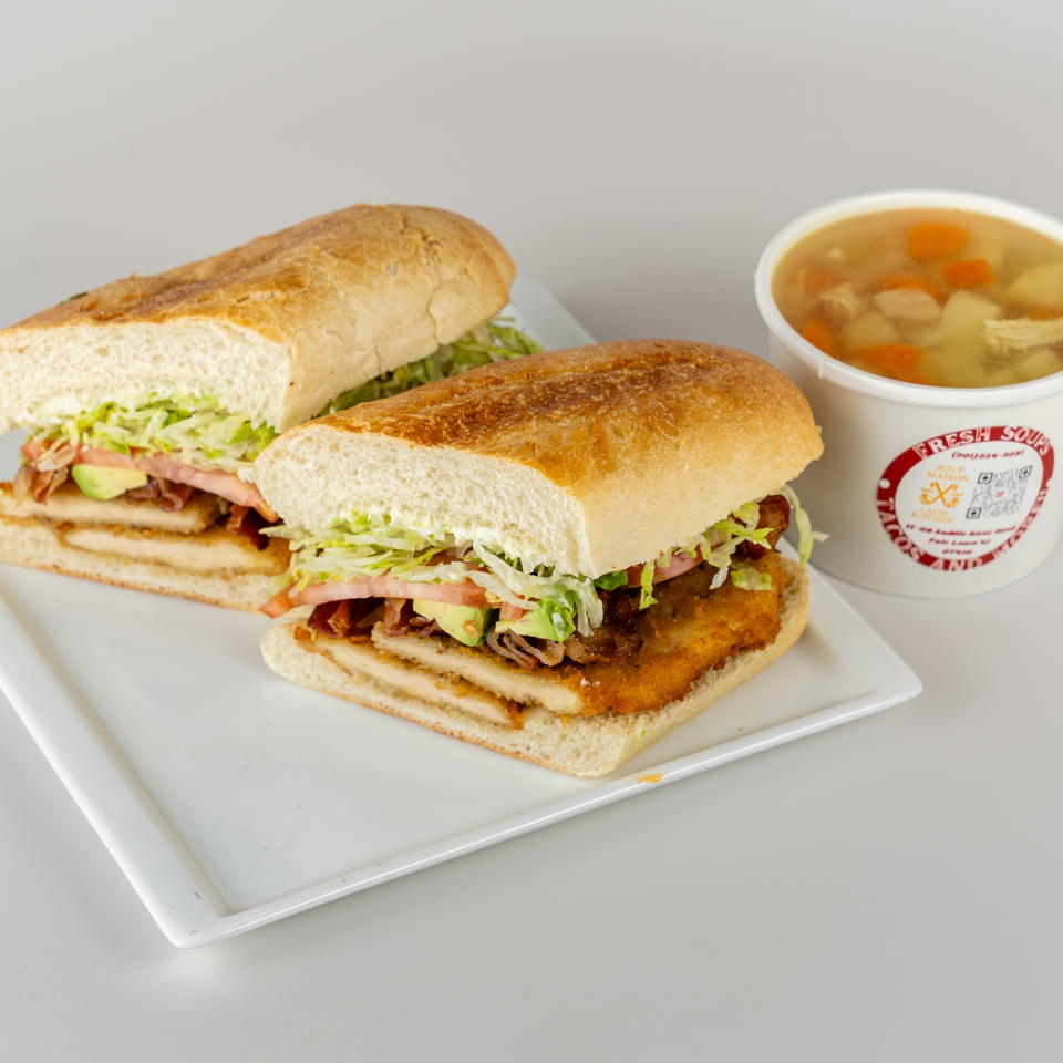 Two halves of a crispy chicken sandwich with lettuce, tomato, and avocado, next to a cup of chicken noodle soup.